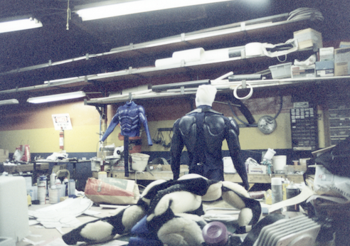 Wide view of the Lowtech workshop with two finished hero suits standing among molds, reference, and benches