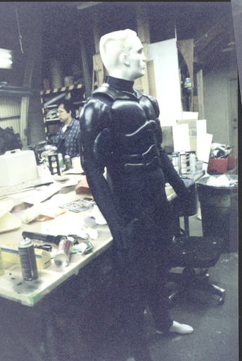 A fully fabricated black hero suit on a mannequin in the Lowtech shop, surrounded by tools and reference